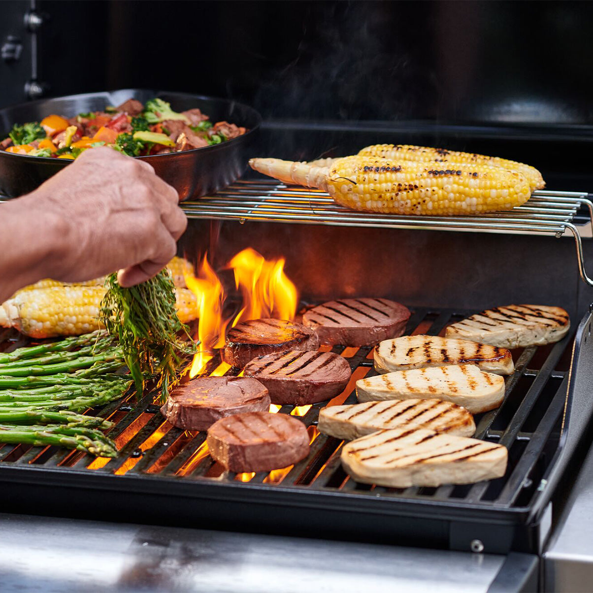 A person grilling steaks with corn and asparagus.